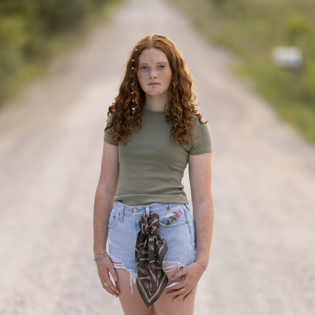 A young person with long curly red hair and freckles stands on a gravel road, wearing a green t-shirt, ripped denim shorts with the Handker Bandanas - No. 116 Edith Bandana by Handker Bandanas tied at the waist, and colorful beads in their hair. Background blurred.