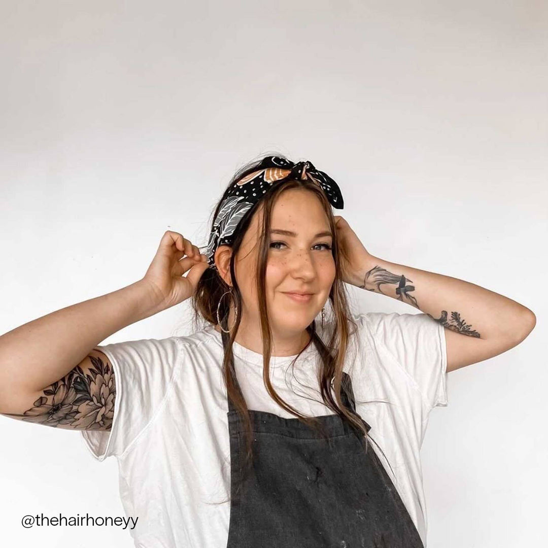A woman with long brown hair smiles at the camera, wearing the Handker Bandanas - No. 046 Amy Bandana by Handker Bandanas, hoop earrings, a white t-shirt, a dark apron, and showcasing floral tattoos on both arms against a plain white background.