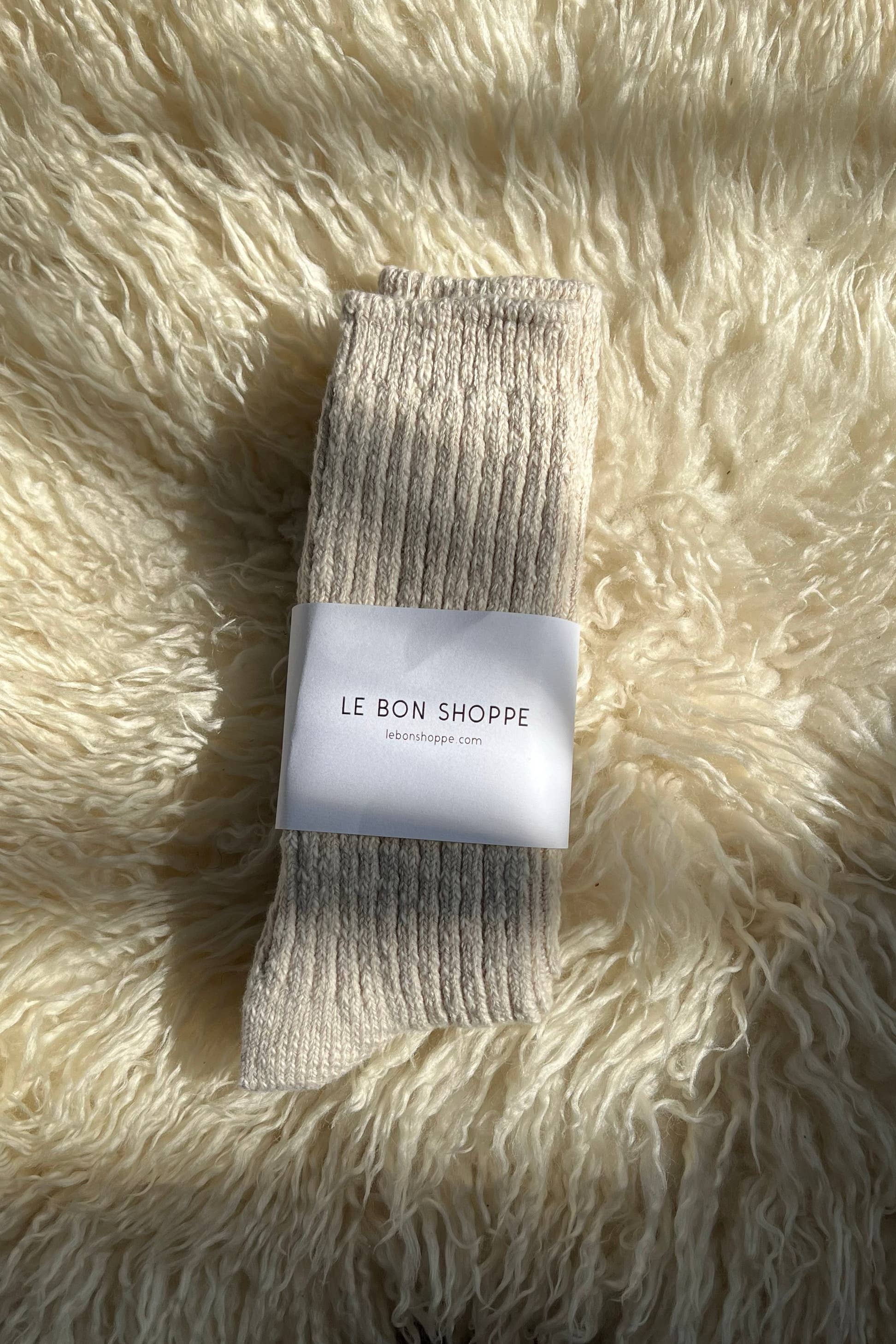 A pair of Le Bon Shoppe Cottage Socks in Oatmeal, wrapped with a branded white label, rests on a fluffy cream surface under natural sunlight.