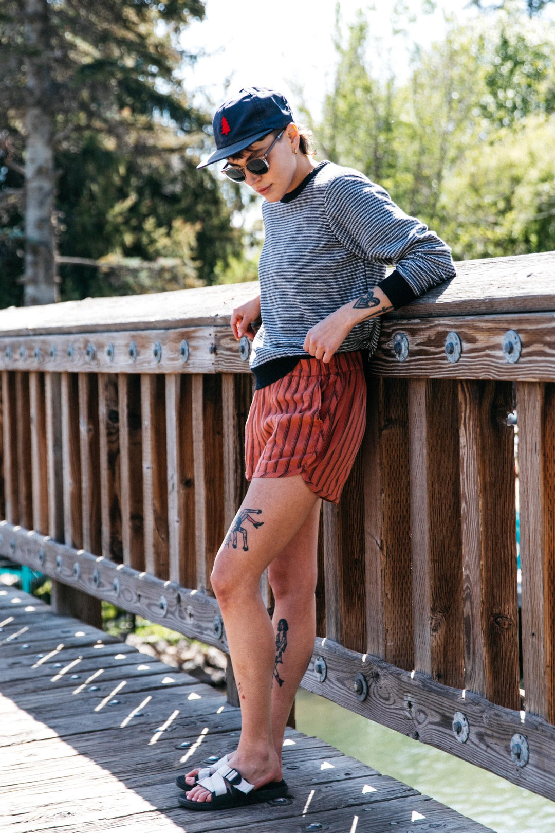 Wearing the Bridge & Burn Ansel Sweater in Charcoal Stripe, this tattooed individual in a cap, sunglasses, and sandals leans on a wooden bridge railing, soaking up the sun amid trees—a true sustainable essential.