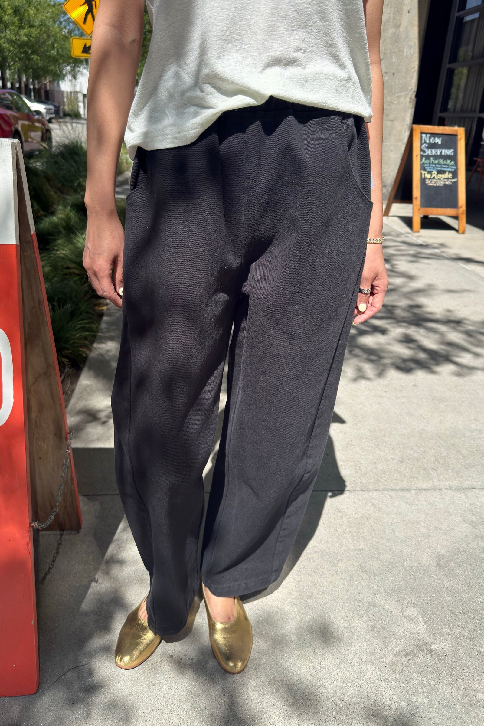 A person stands on a sidewalk wearing the Le Bon Shoppe - Arc Pant in black, paired with a light-colored top and gold shoes. Sunlight and shadows fall across the ground, with a red sign, greenery, and a chalkboard sign visible in the background.