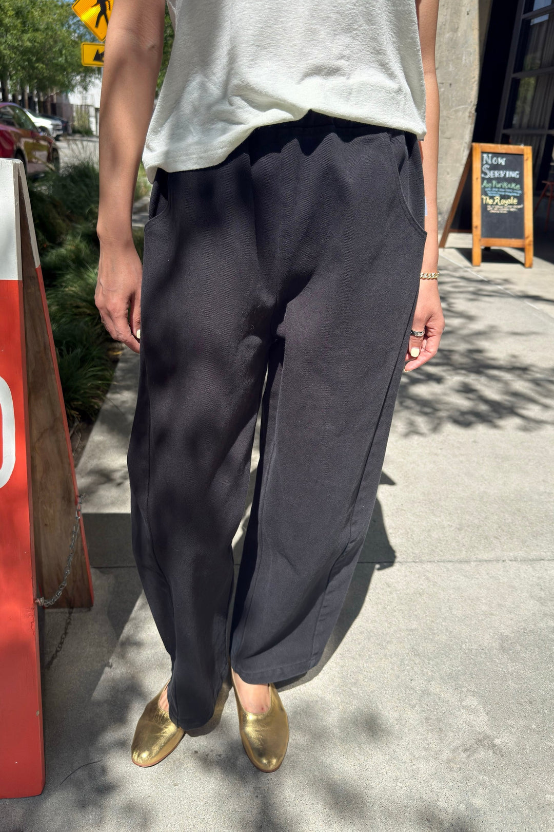 A person stands on a sidewalk wearing the Le Bon Shoppe - Arc Pant in black, paired with a light-colored top and gold shoes. Sunlight and shadows fall across the ground, with a red sign, greenery, and a chalkboard sign visible in the background.