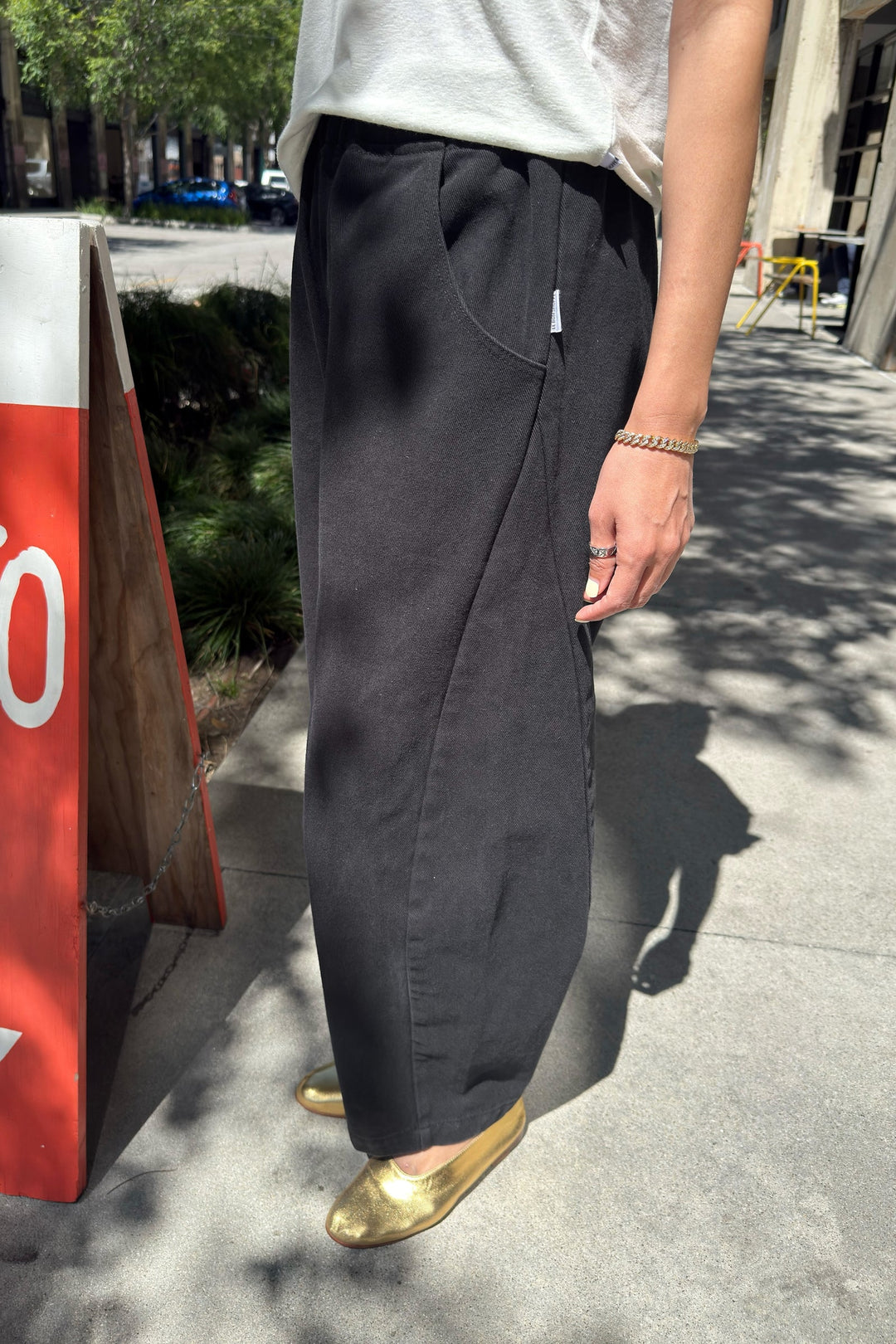 A person stands on a sidewalk wearing Le Bon Shoppe - Arc Pant in Black, paired with a white shirt, a gold bracelet, and gold shoes. The sun casts shadows on the pavement while a red and white sign is partially visible in the background.