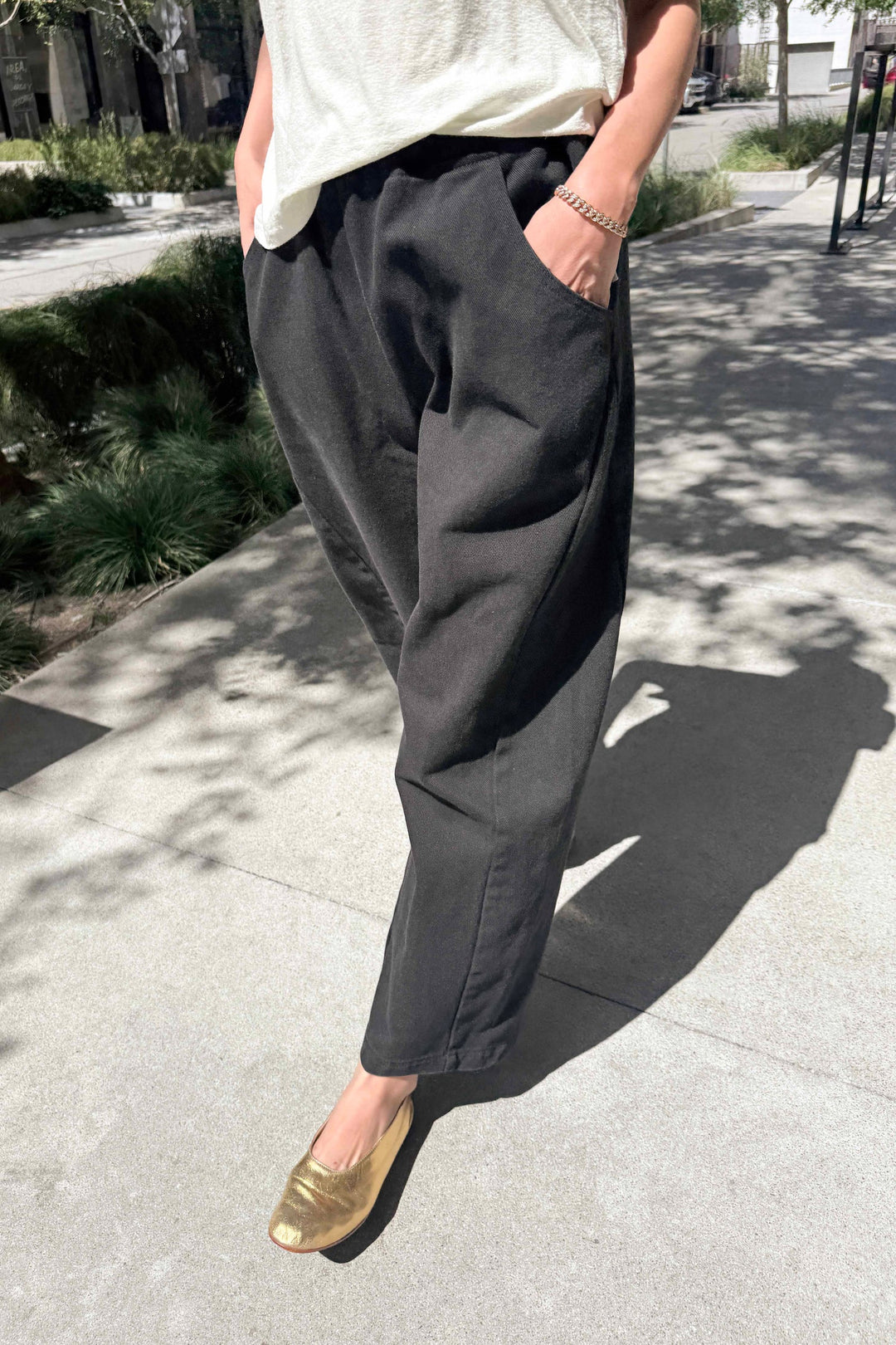 A person stands outdoors in the sunlight with their hands in the pockets of Le Bon Shoppe's Arc Pant in Black, paired with a white top and shiny gold shoes. Only the lower half of their body is visible.