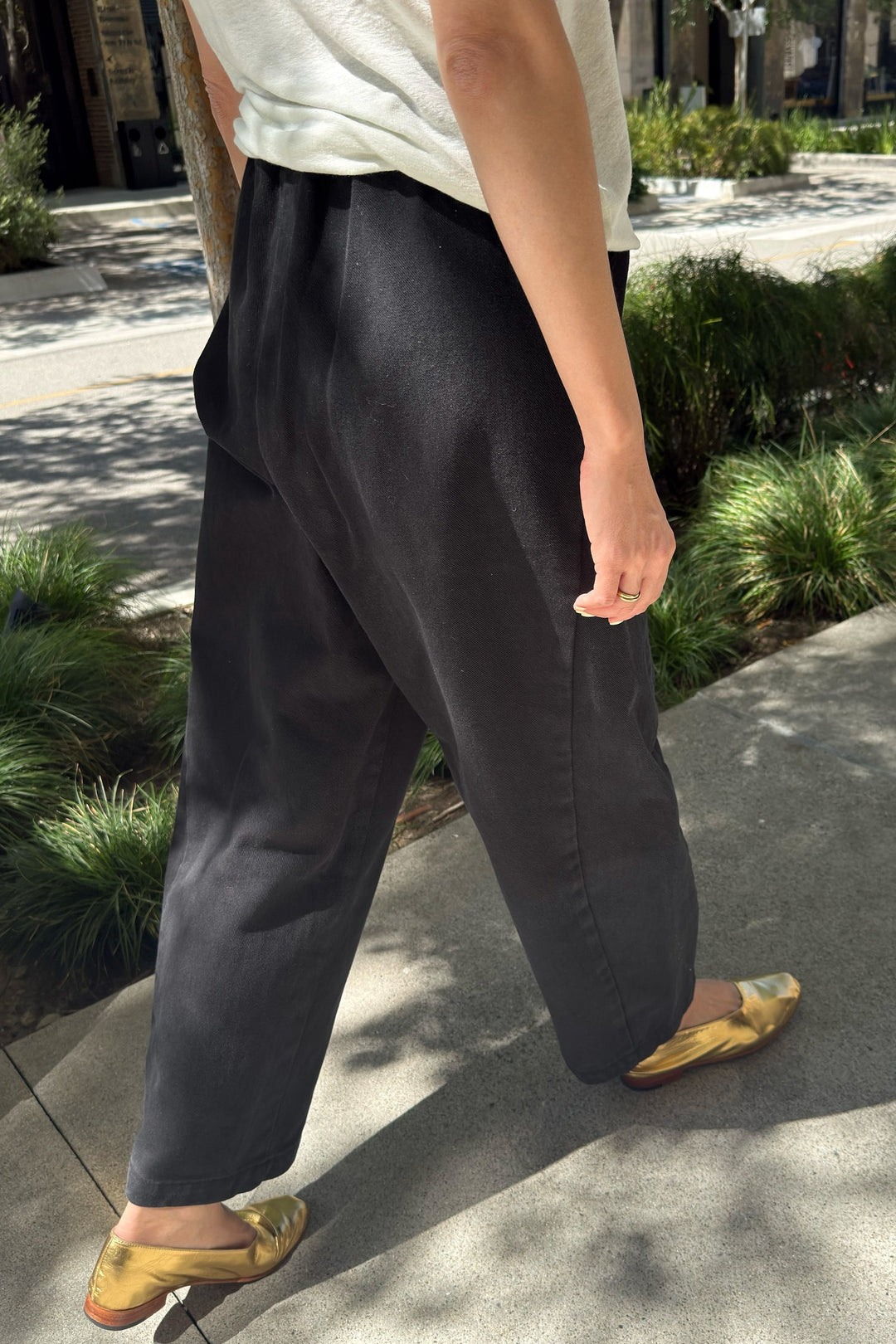 A person is seen from the waist down wearing Le Bon Shoppe - Arc Pant in Black, paired with a white shirt and gold shoes as they walk along a sidewalk bordered by green bushes on a sunny day.