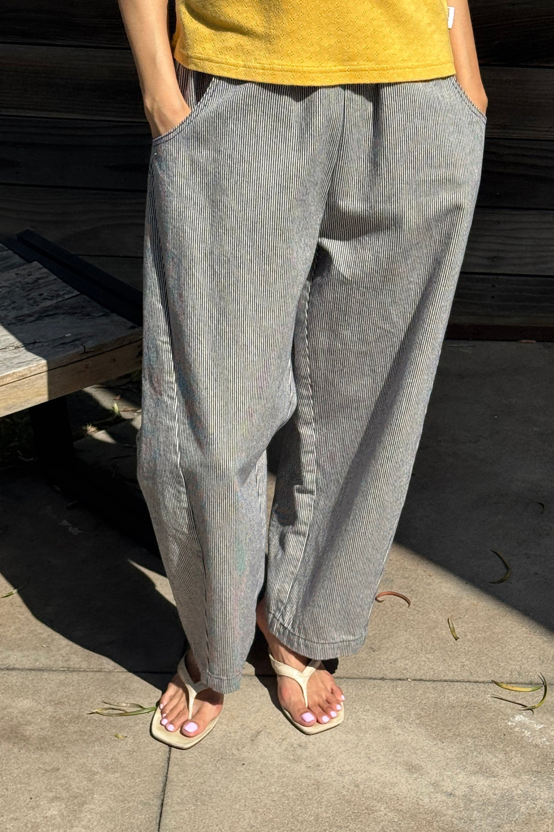 A person wearing Le Bon Shoppe - Arc Pant in Blue Stripe, paired with a yellow shirt and white sandals, stands outdoors on a sunny day with hands in pockets; only the lower body is visible.