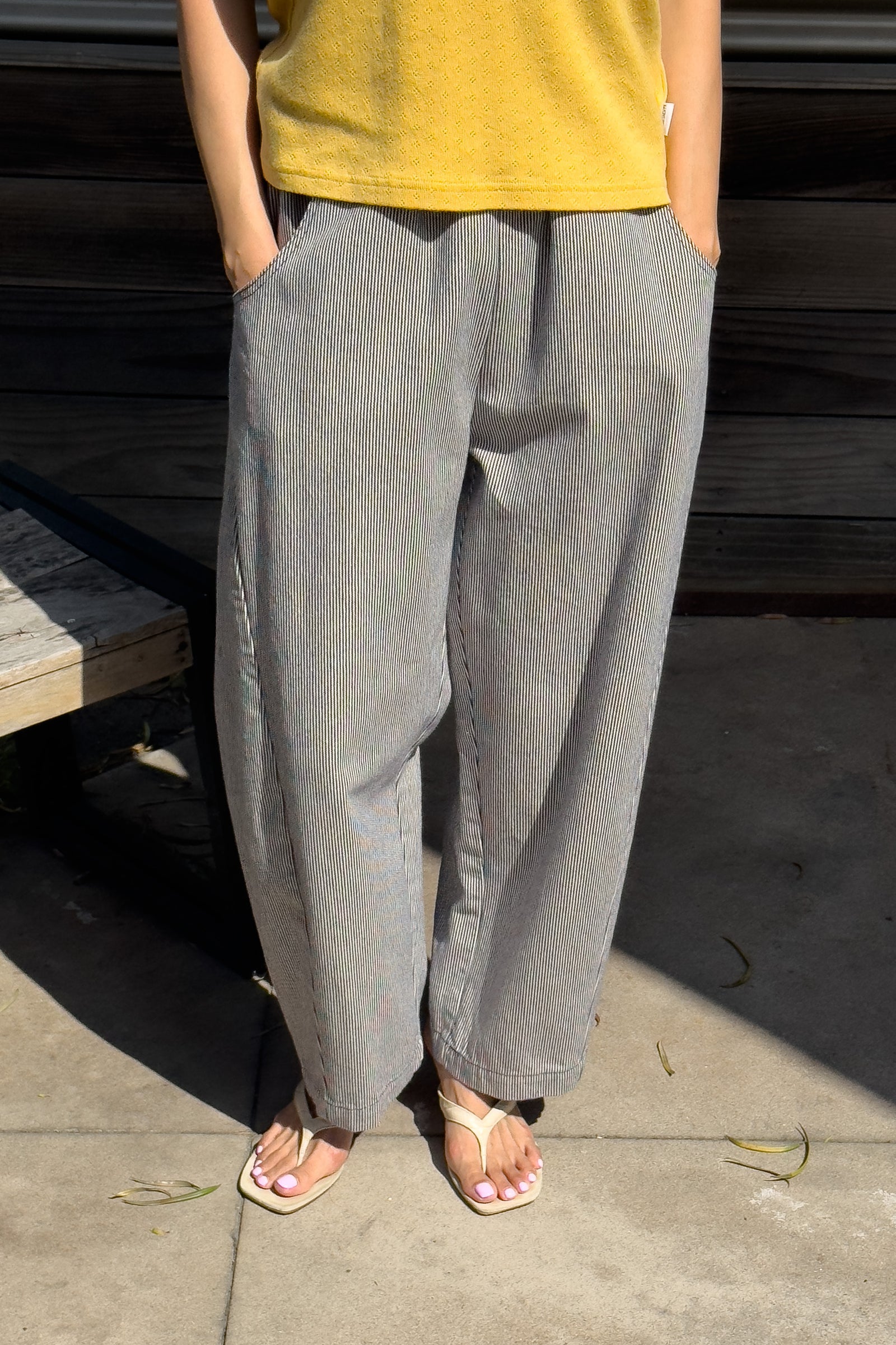 A person stands outdoors wearing a yellow shirt, Le Bon Shoppe - Arc Pant in Blue Stripe with hands in pockets, and white sandals. Only their lower body is visible.