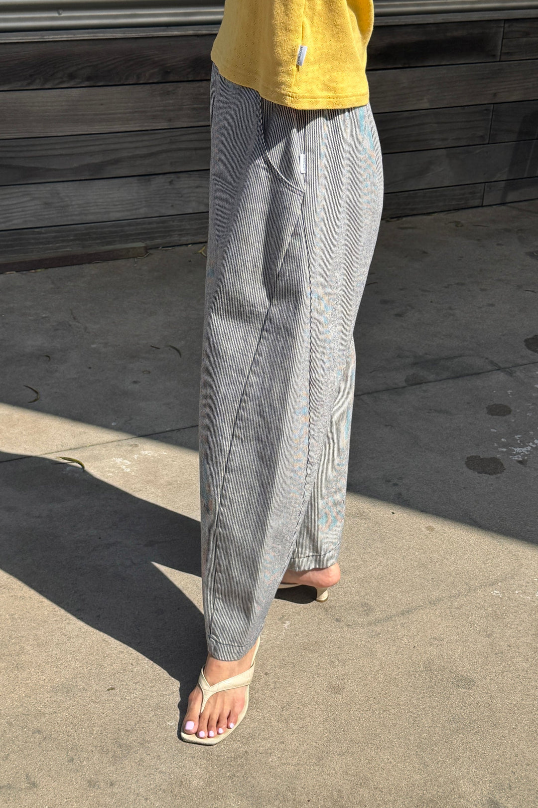 A person wearing a yellow shirt, Le Bon Shoppe Arc Pant in Blue Stripe, and open-toed heels stands on a sunny sidewalk near a wooden wall.