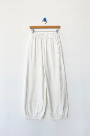 The Le Bon Shoppe Balloon Pant in white cotton features a loose, balloon leg fit with an elastic waistband and cuffs, displayed on a wooden hanger against a plain white wall.