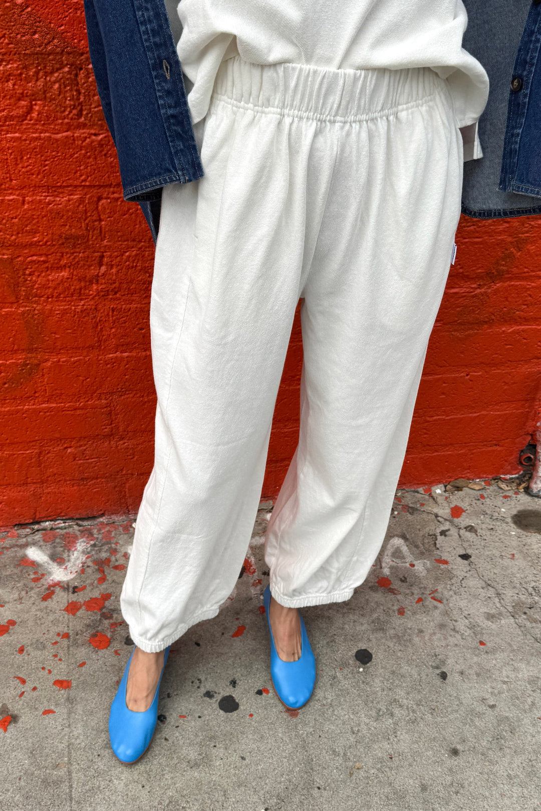 Wearing Le Bon Shoppe Balloon Pant in White Cotton, a cream shirt, and a blue denim jacket, the person stands by a bright red brick wall in blue ballet flats on a gray, paint-splattered sidewalk.