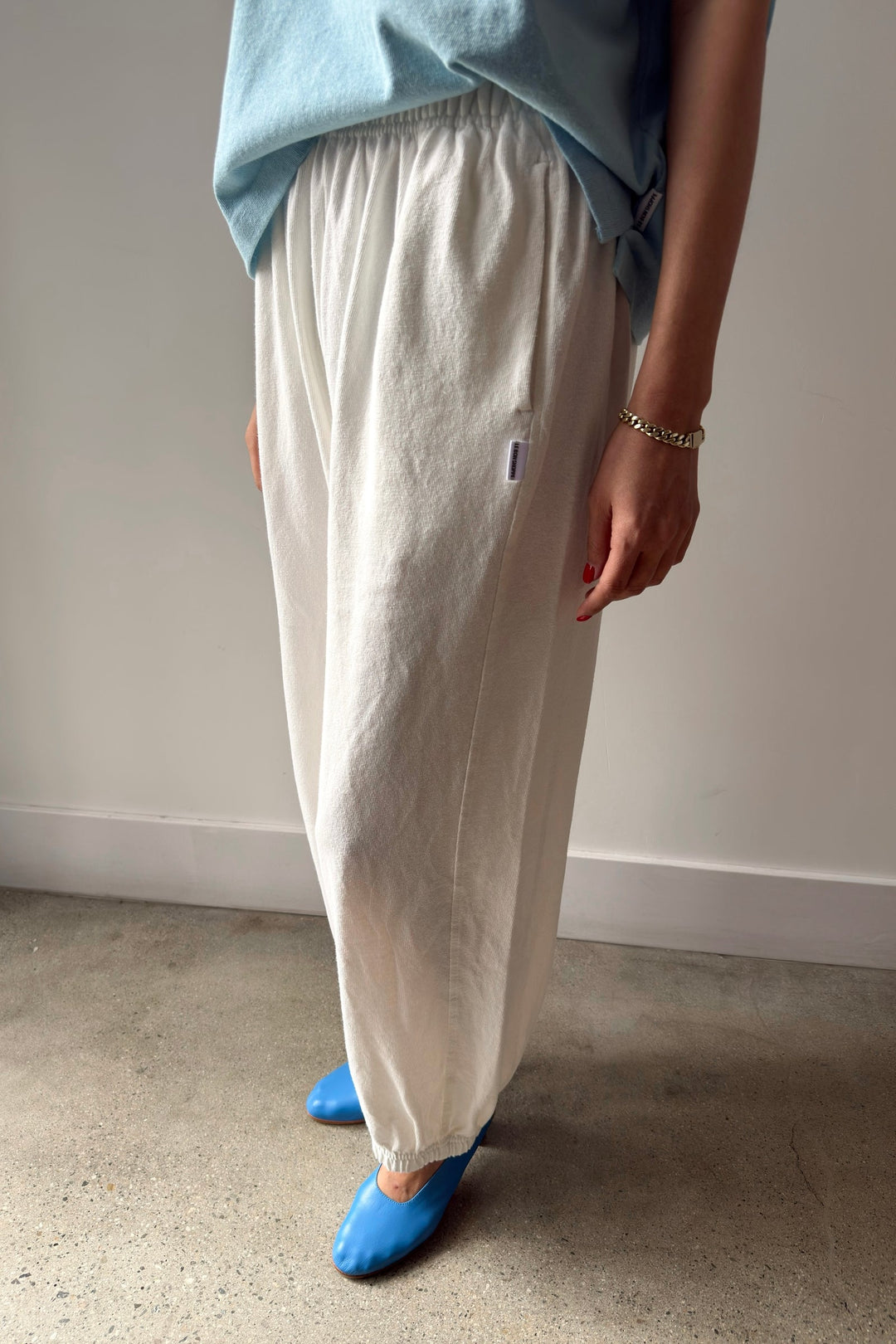 A person stands indoors on a polished concrete floor, wearing Le Bon Shoppe Balloon Pant in white cotton, a light blue top, bright blue shoes, and a gold bracelet. Their right hand is relaxed at their side.