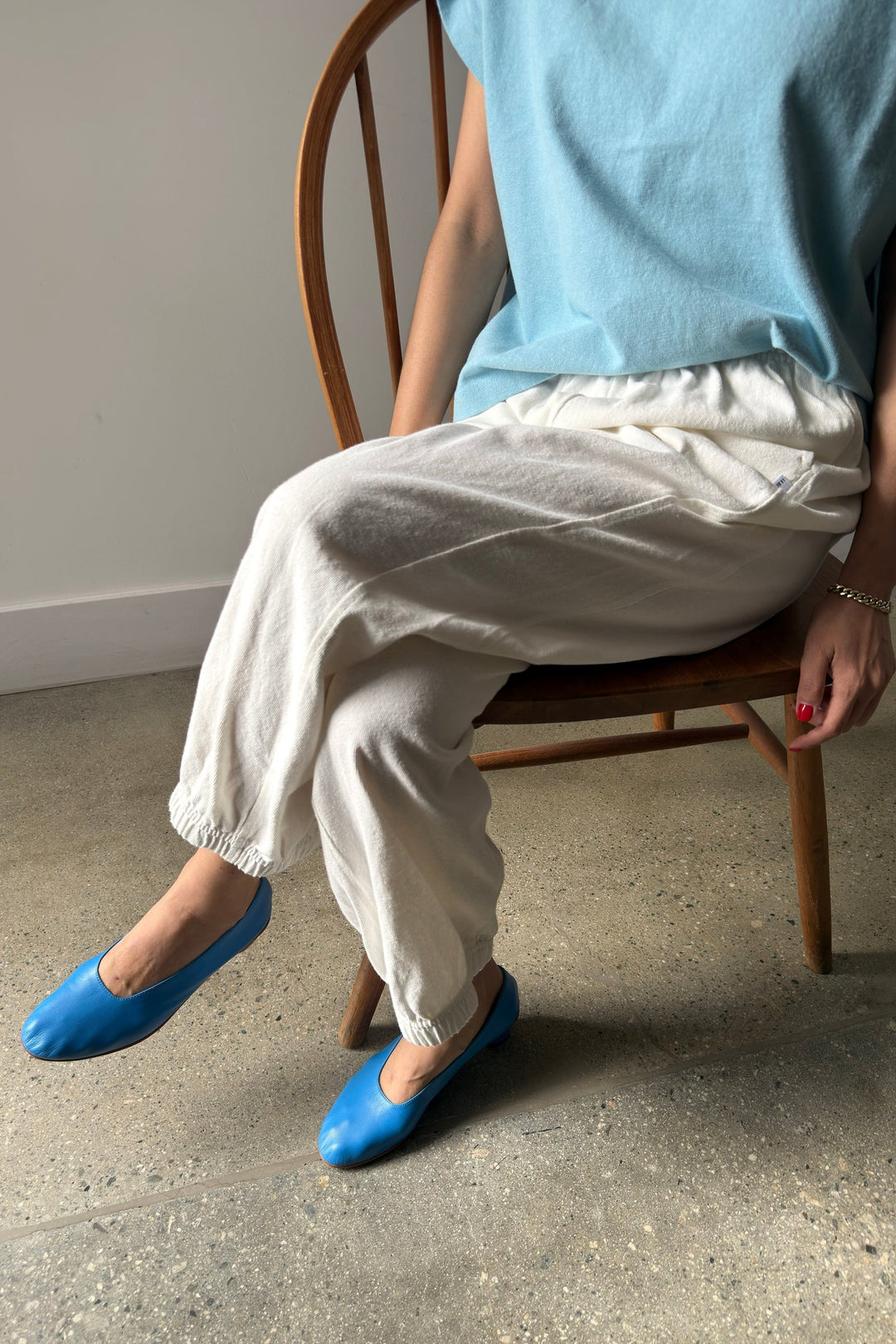 A person sits on a wooden chair wearing Le Bon Shoppe - Balloon Pant / White Cotton. Only their lower body and part of an arm are visible. The floor is concrete, and the background is plain.