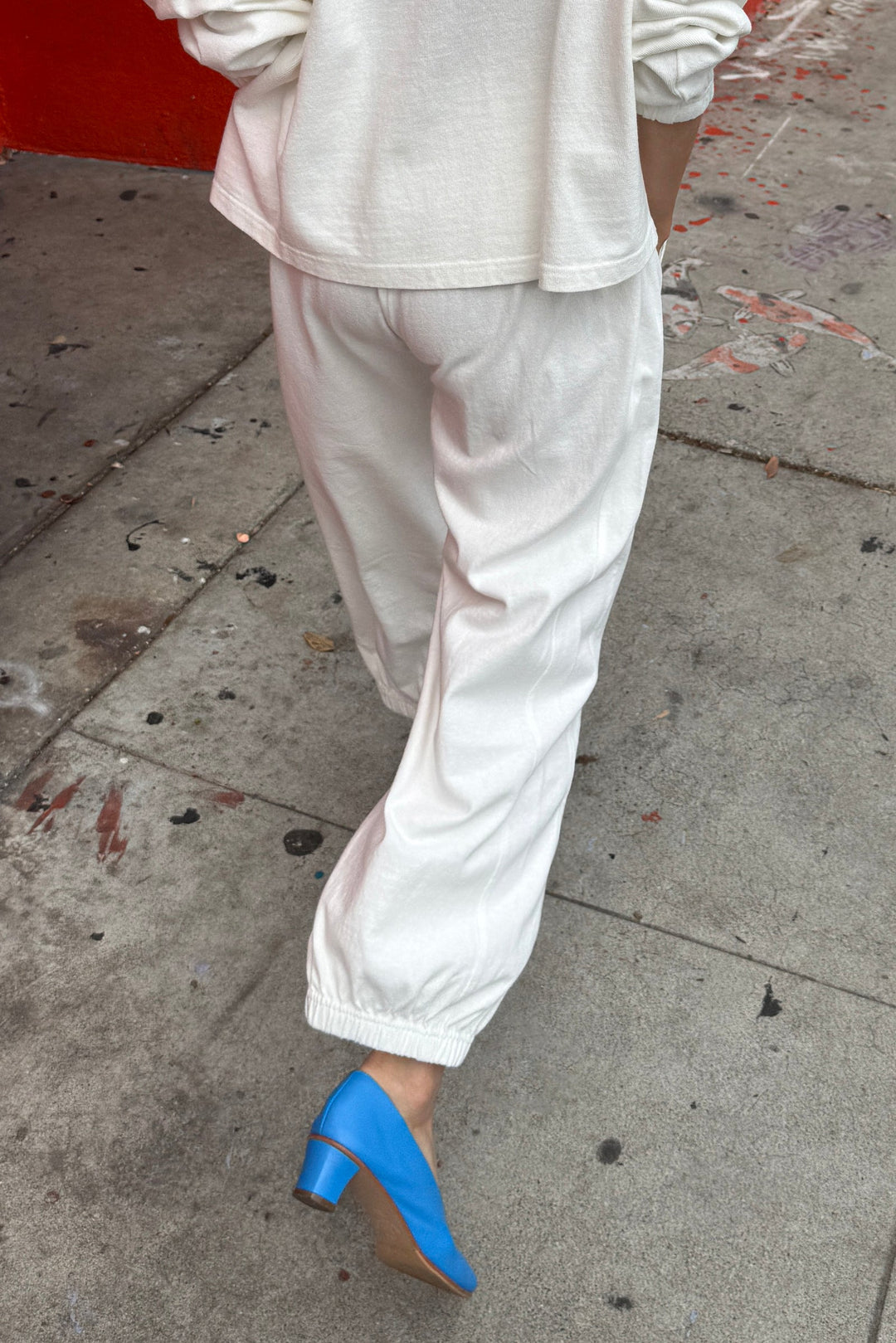 A person walks on a city sidewalk wearing Le Bon Shoppe's Balloon Pant in white cotton with a matching long-sleeve top and bright blue heeled shoes. The ground is stained and littered, with a red wall to the left.