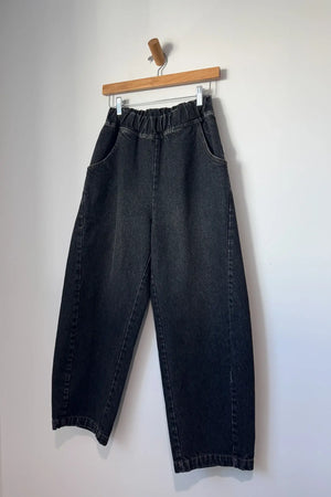 The Le Bon Shoppe Arc Pant in black denim, crafted from heavy-weight fabric with a comfortable elastic waistband and wide-leg silhouette, hangs on a wooden hanger against a plain white wall.