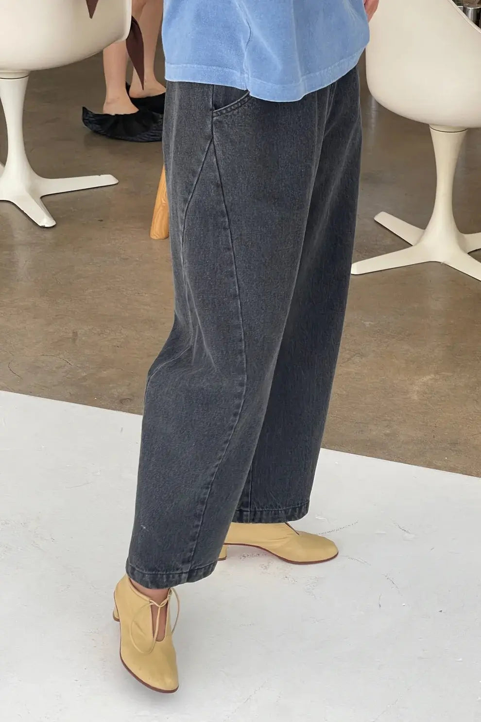A person is shown from the waist down wearing Le Bon Shoppe - Arc Pant in Black Denim, styled with beige heeled shoes. The cropped, loose-fitting jeans are made from heavy-weight denim and stand out against a white floor, while other people and white chairs can be seen in the background on a light brown floor.