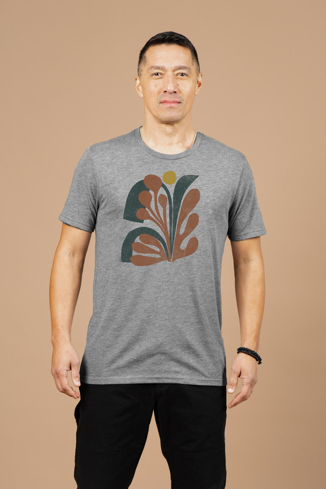 A man with short dark hair smiles slightly against a beige background, wearing the Bridge & Burn Men's Bloom Tee in grey—an organic cotton t-shirt featuring a colorful abstract design with curved shapes and a small yellow circle.