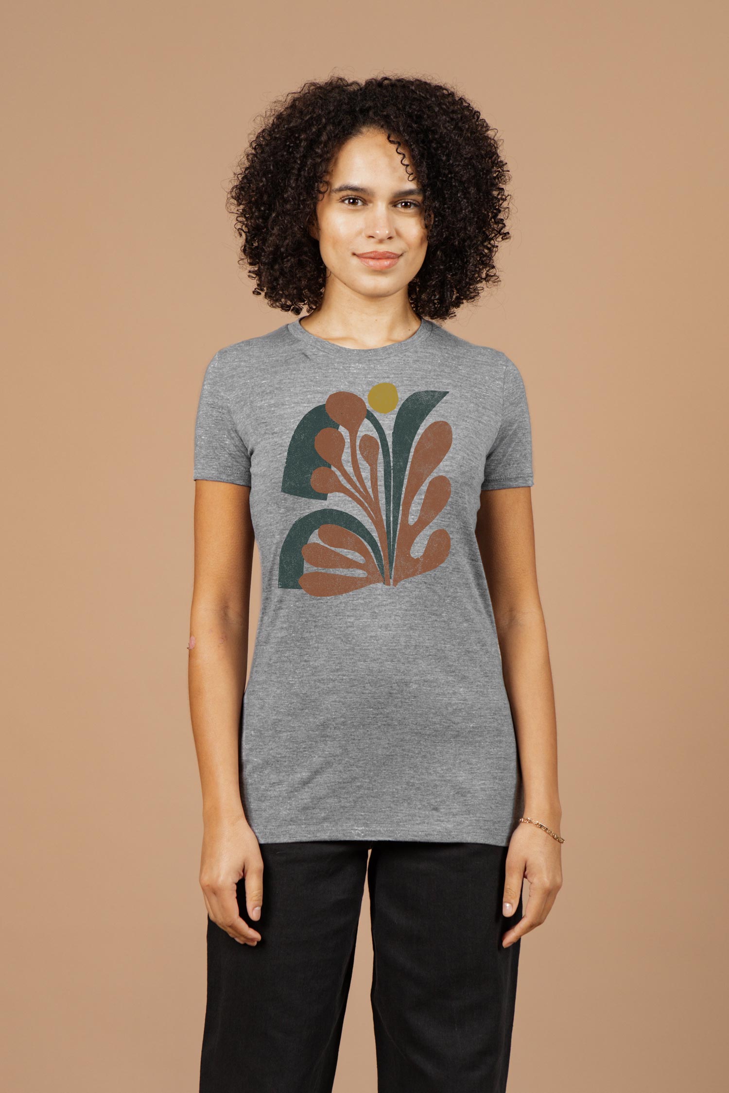 A woman with curly hair wears the Bridge & Burn Women's Bloom Tee in grey, featuring a colorful floral design, standing against a plain beige background.