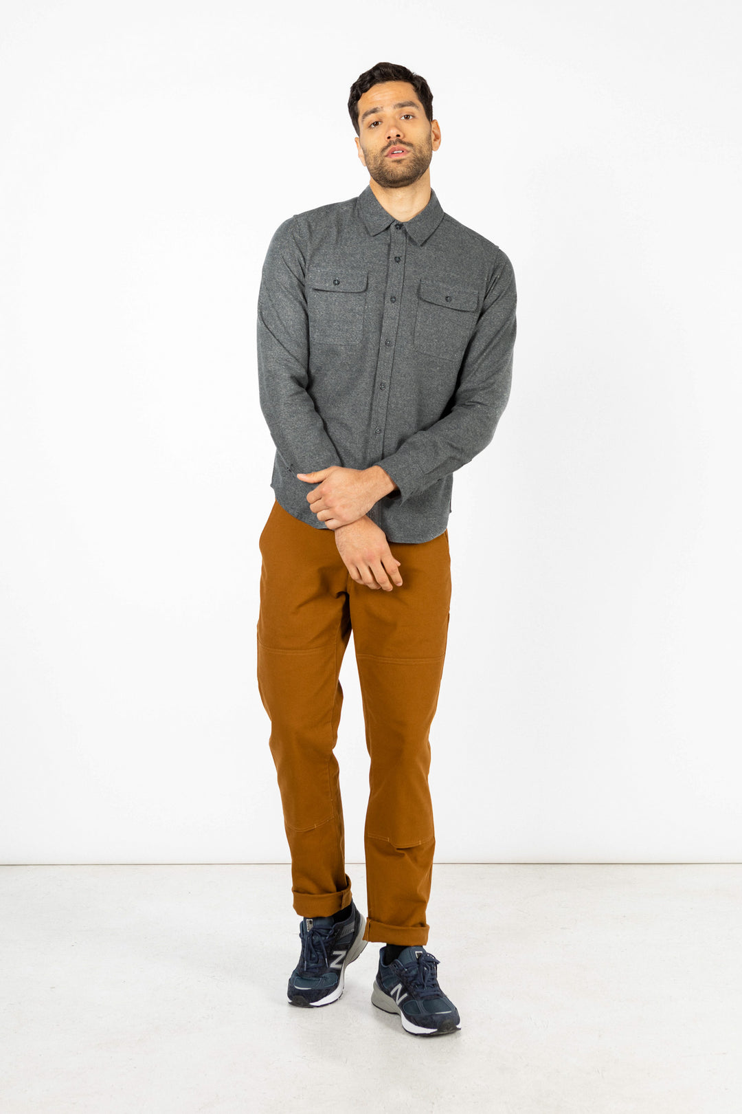 A man stands against a plain white background, wearing the Bridge & Burn Cole Slim Shirt in Stone, brown pants, and navy blue sneakers. He has a neutral expression and his arms are loosely crossed in front of him.