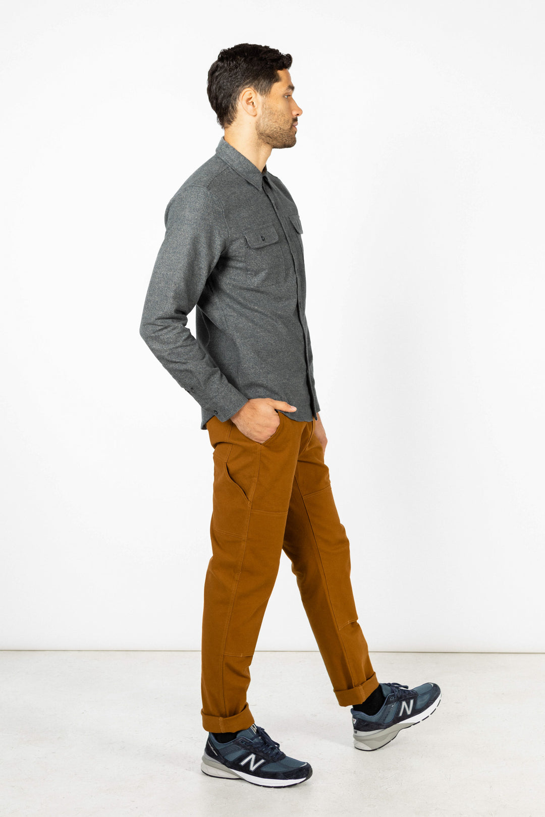 A man in the Bridge & Burn Cole Slim Shirt in Stone, brown pants, and navy New Balance sneakers stands in profile against a plain white background.