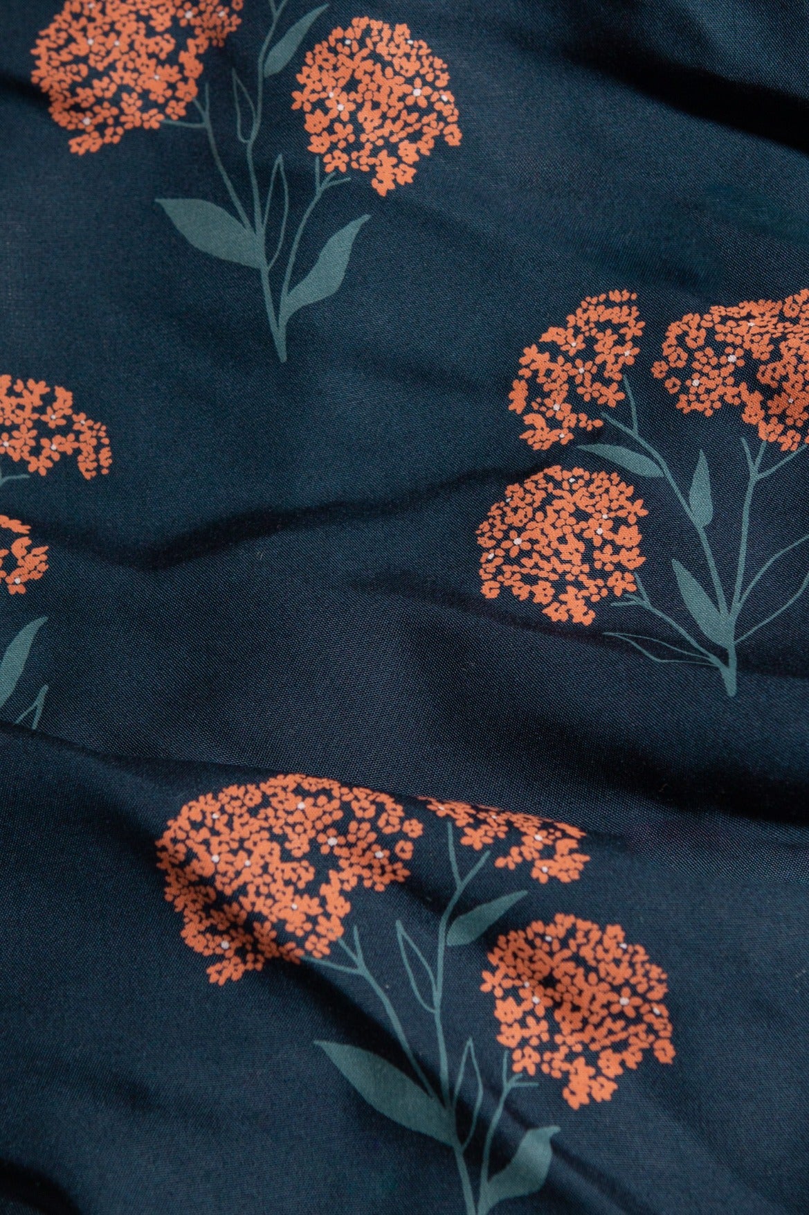 Close-up of the dark blue Ira Dress / Wildflower by Bridge & Burn, showcasing orange clustered floral patterns with green stems and leaves on sustainable fabric, gently folded for a textured look.