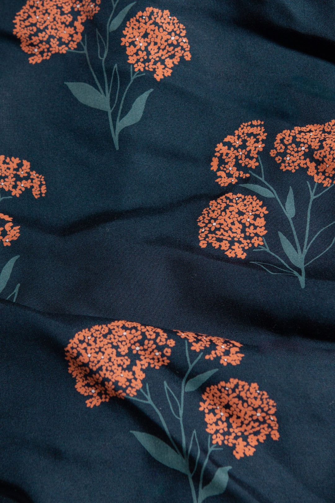 Close-up of the dark blue Ira Dress / Wildflower by Bridge & Burn, showcasing orange clustered floral patterns with green stems and leaves on sustainable fabric, gently folded for a textured look.