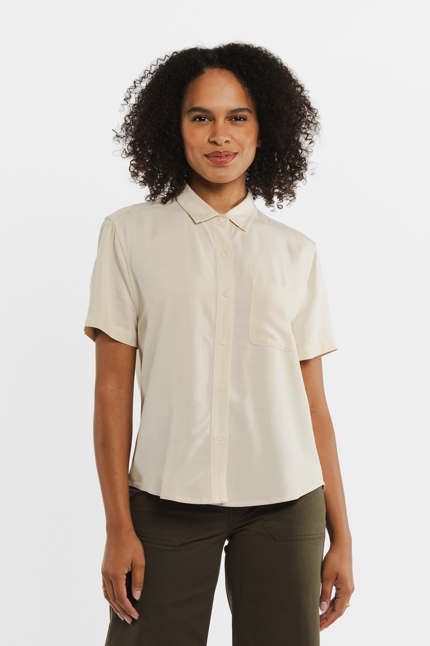 A person with curly hair wears the Bridge & Burn Greer Cropped Shirt in Cream and dark green pants, standing and smiling against a plain white background.