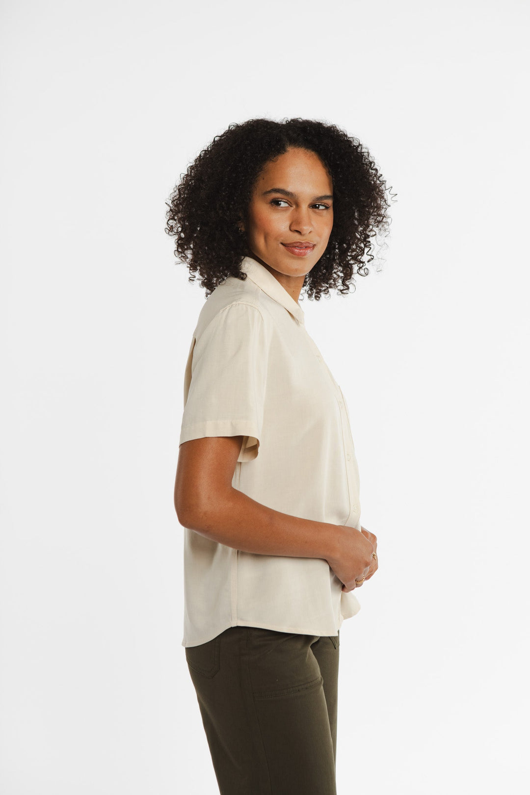 A woman with curly hair, wearing the Bridge & Burn Greer Cropped Shirt in cream and dark pants, stands in profile against a plain white background, smiling softly.