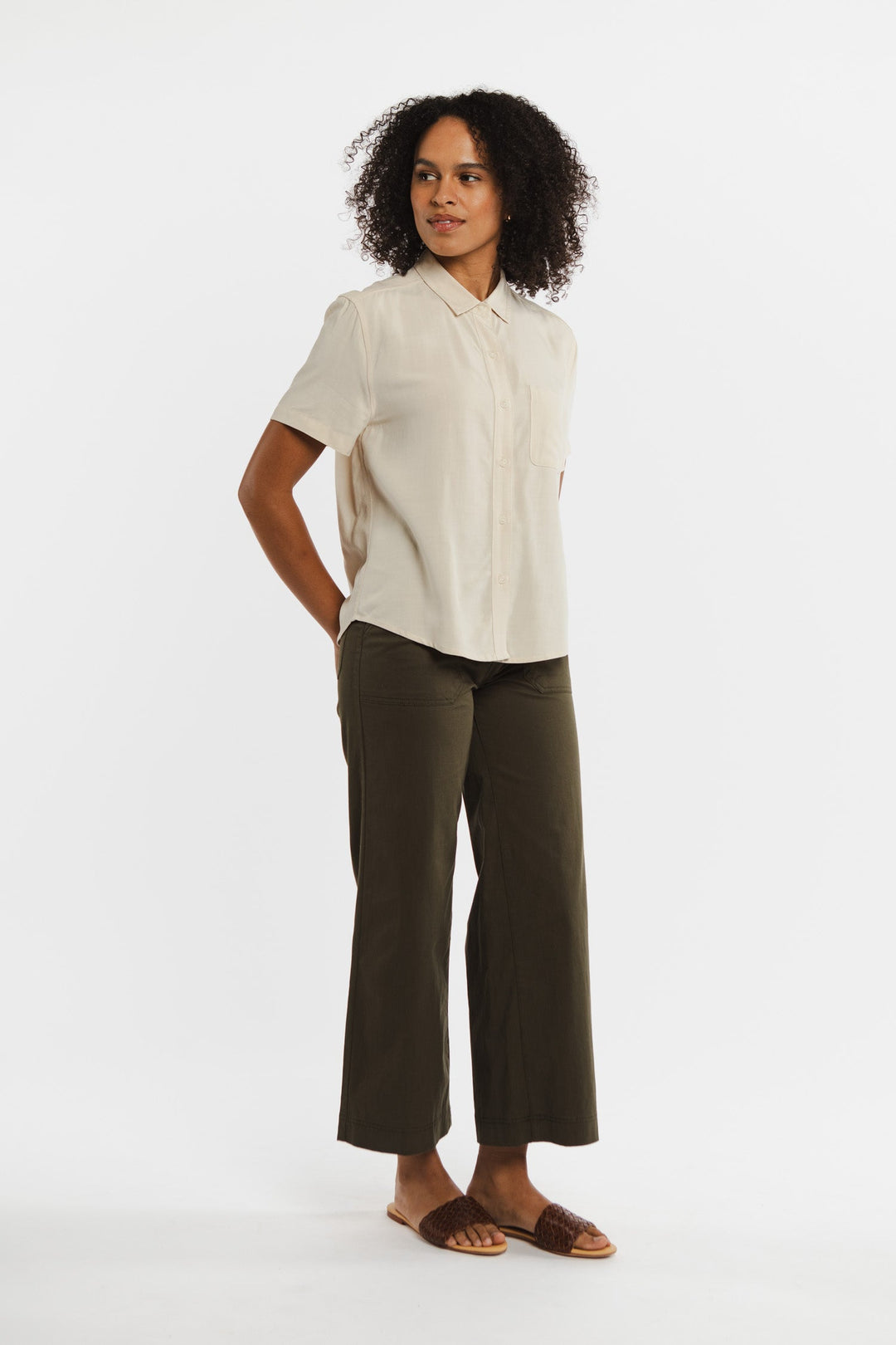 A person with curly hair stands against a white background, wearing the Bridge & Burn Greer Cropped Shirt in cream, paired with olive green wide-leg pants and brown sandals. Their hands are behind their back as they look slightly to the side.