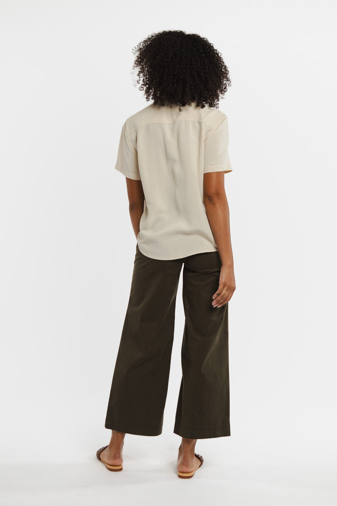 A person with curly hair stands facing away, wearing the Bridge & Burn Greer Cropped Shirt in Cream, wide-leg dark olive pants, and flat sandals against a plain white background.