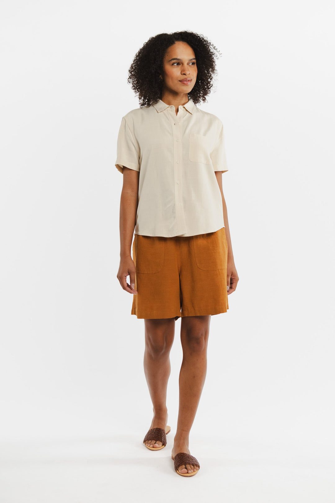 A woman with curly hair and a relaxed expression stands against a white background, wearing the Bridge & Burn Greer Cropped Shirt in Cream, high-waisted burnt orange twill shorts, and brown slide sandals.