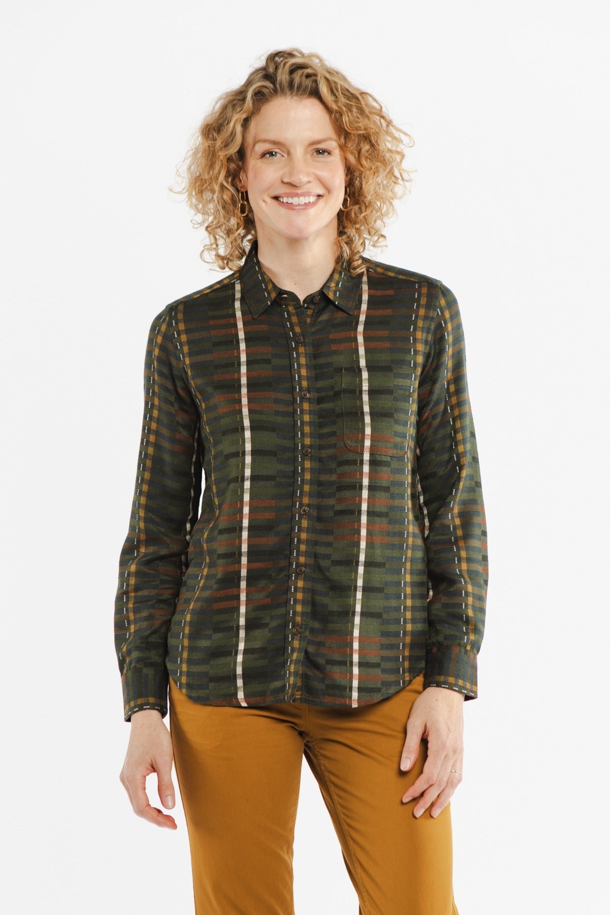 A woman with curly blonde hair smiles, wearing the Bridge & Burn Ash Slim Shirt in Olive Stripe and mustard pants, standing against a plain white background.