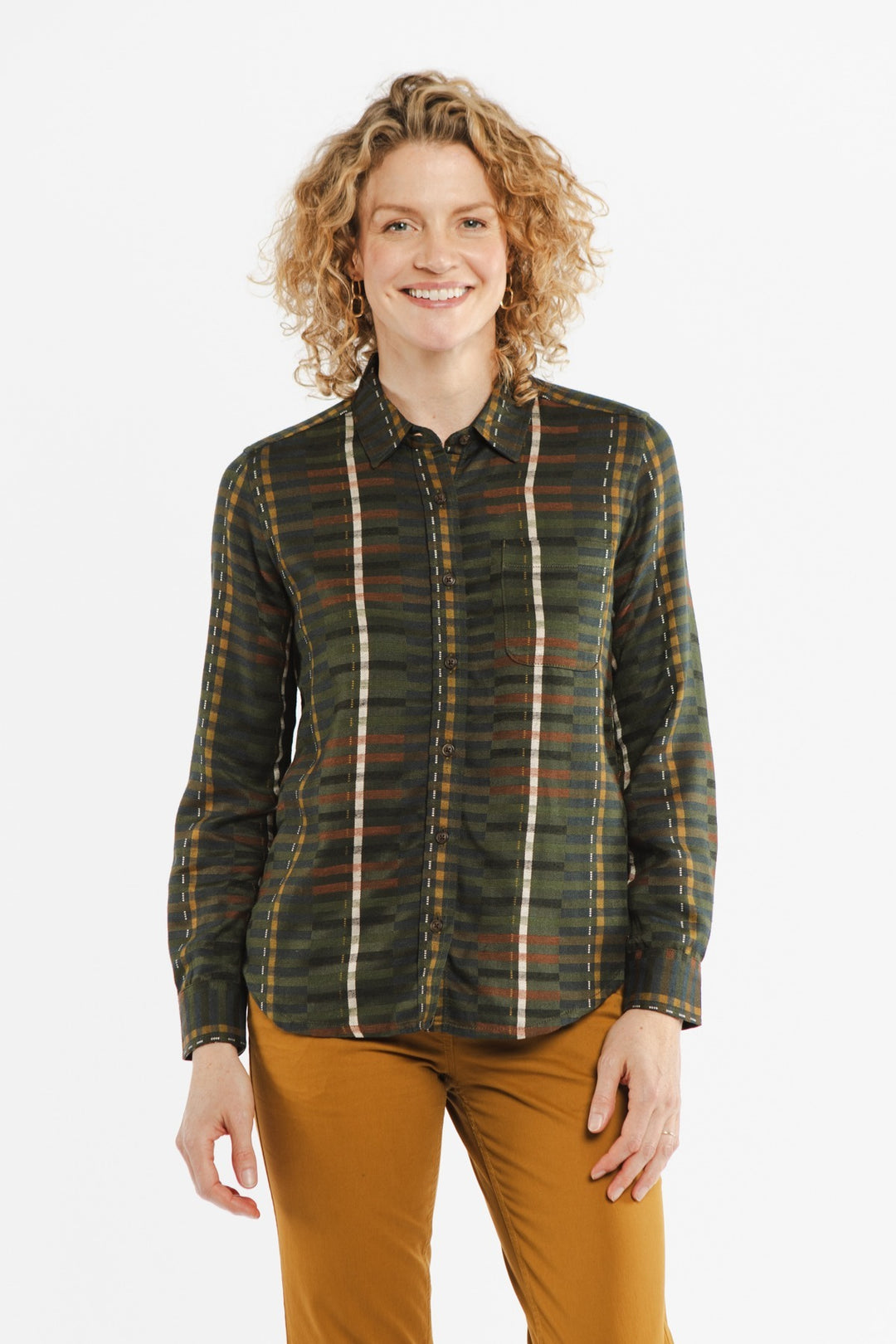 A woman with curly blonde hair smiles, wearing the Bridge & Burn Ash Slim Shirt in Olive Stripe and mustard pants, standing against a plain white background.