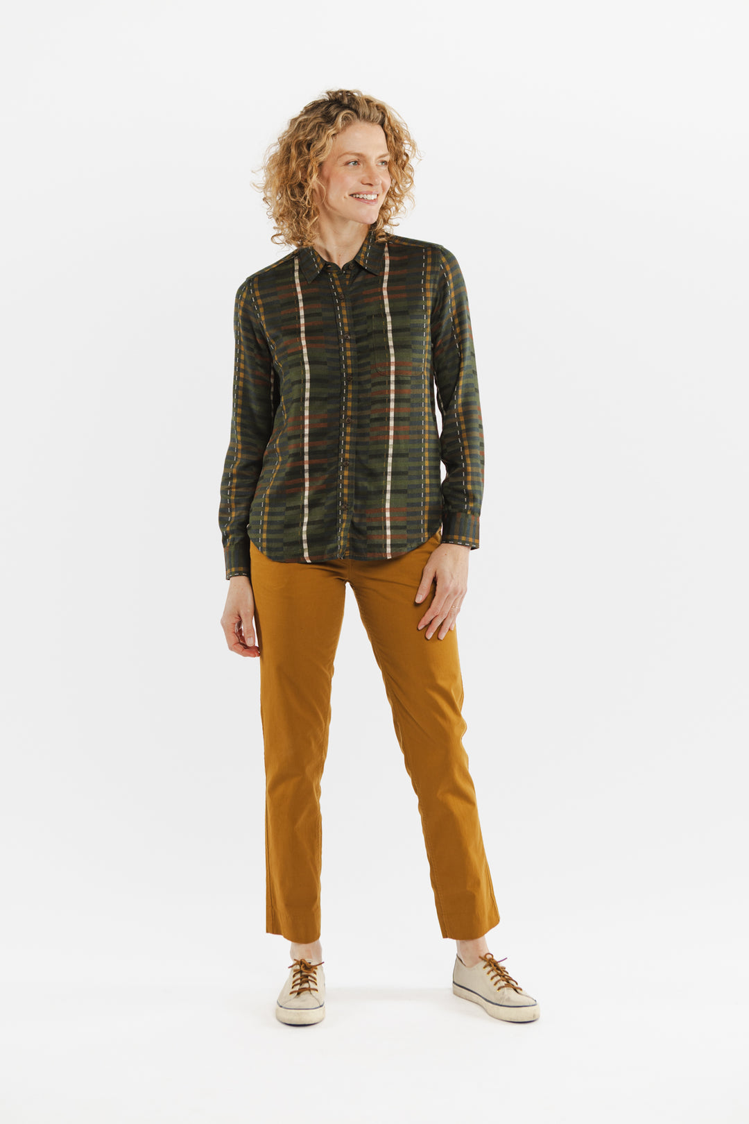 A person with curly hair stands smiling, wearing the Bridge & Burn Ash Slim Shirt in Olive Stripe, paired with mustard yellow pants and beige sneakers against a plain white background.