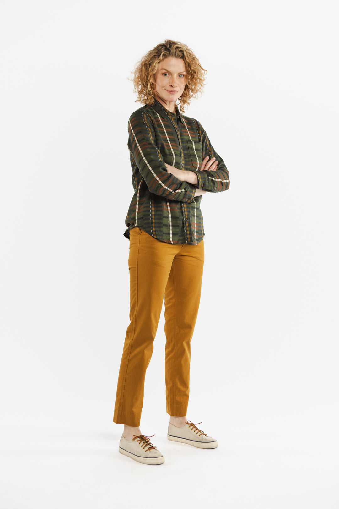 A person with curly blond hair stands confidently with arms crossed, wearing the Bridge & Burn Ash Slim Shirt in Olive Stripe, mustard yellow pants, and white sneakers against a plain white background.