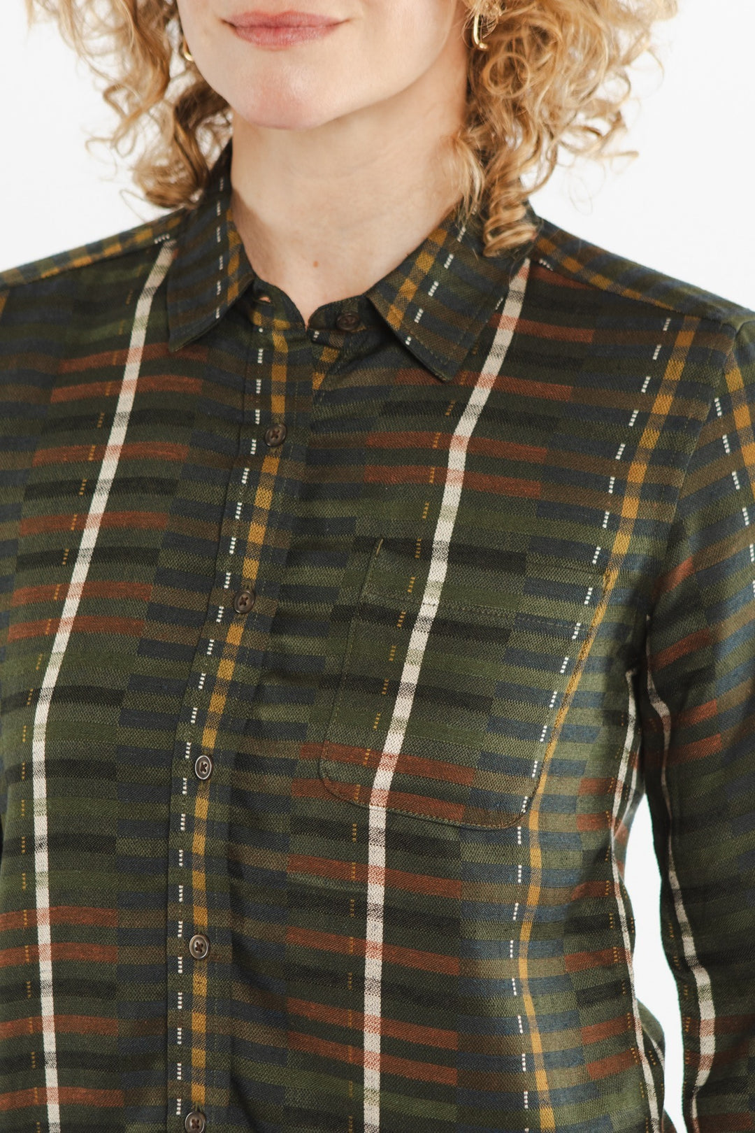 A woman with curly blonde hair models the Bridge & Burn Ash Slim Shirt in Olive Stripe, featuring a green, brown, and yellow plaid pattern with olive stripes and a small chest pocket. Only her lower face and upper torso are visible.