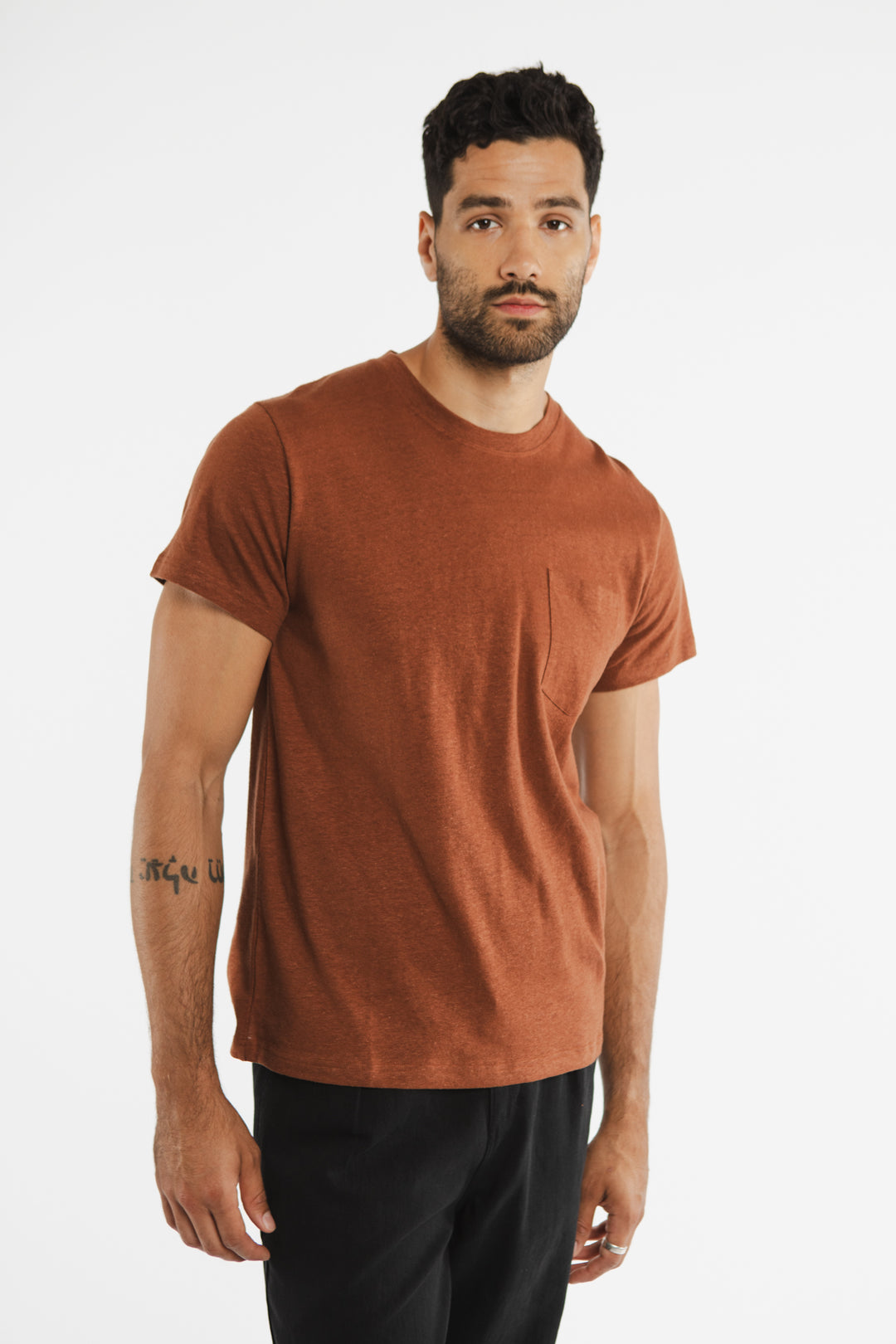 A man with short dark hair and a beard wears the Bridge & Burn Organic Hemp Pocket Tee in Burnt Umber with black pants, standing against a plain white background and looking slightly toward the camera.