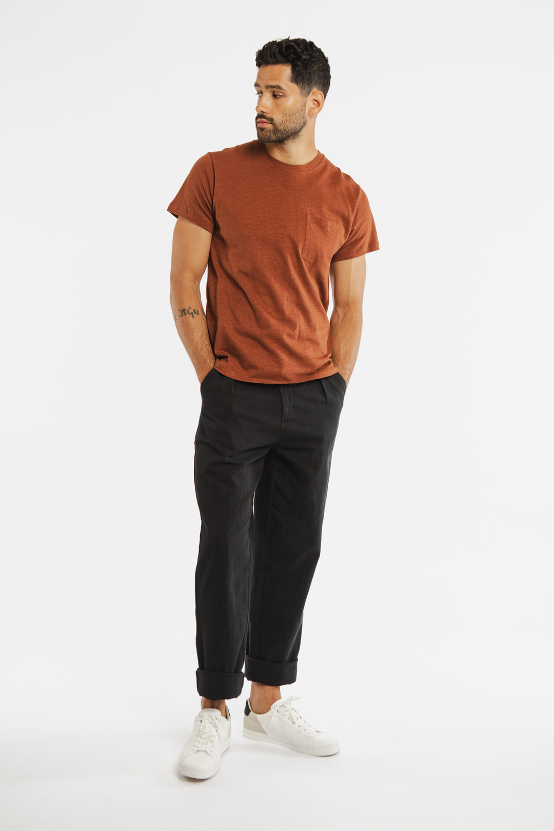 A man with short dark hair, a beard, and a left forearm tattoo stands against a white background wearing Bridge & Burn's Organic Hemp Pocket Tee in Burnt Umber, black cuffed pants, and white sneakers. His hands are in his pockets.