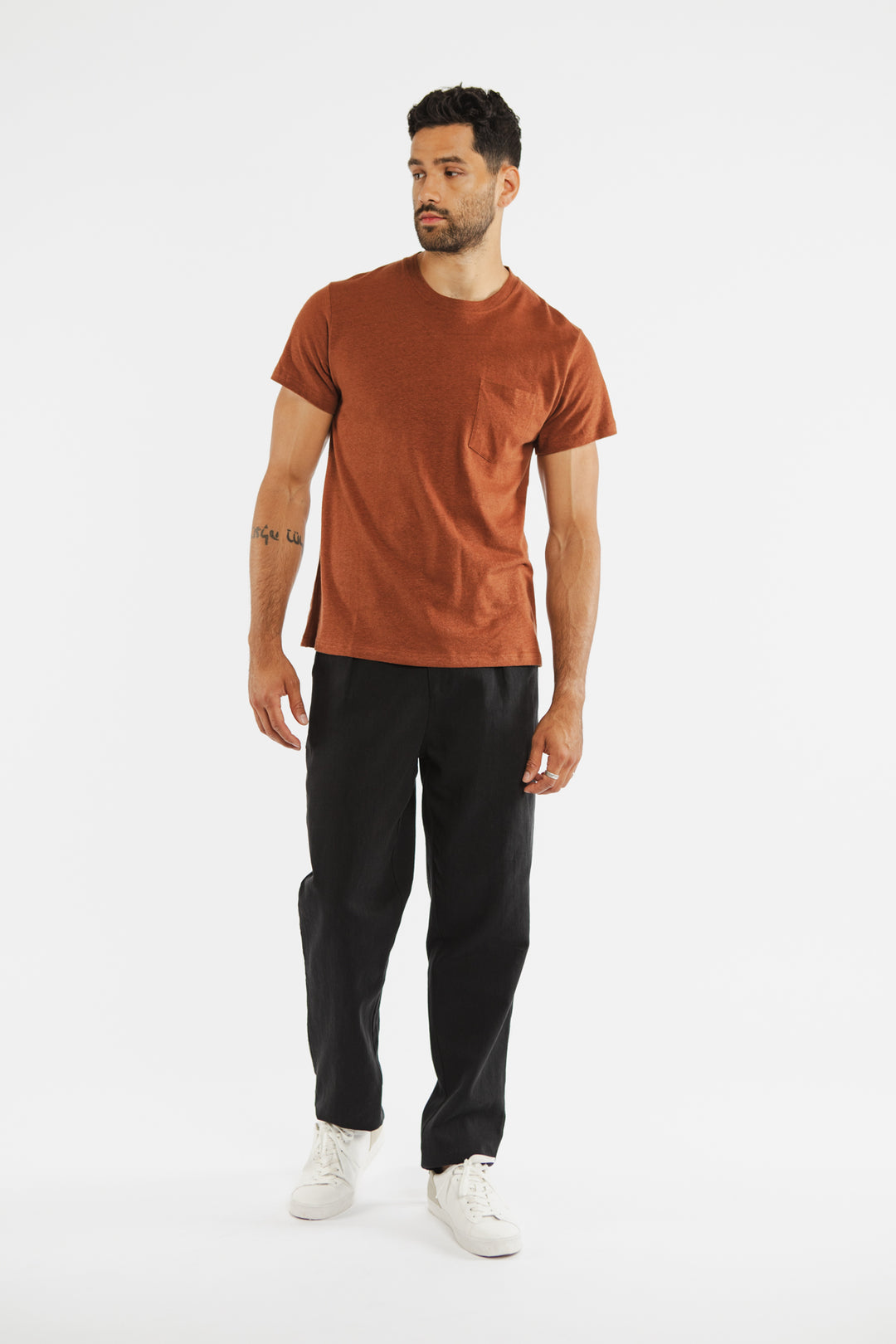 A man stands against a white background wearing the Bridge & Burn Organic Hemp Pocket Tee in Burnt Umber, paired with black pants and white sneakers. He has short dark hair, a beard, and looks slightly left with one hand by his side.