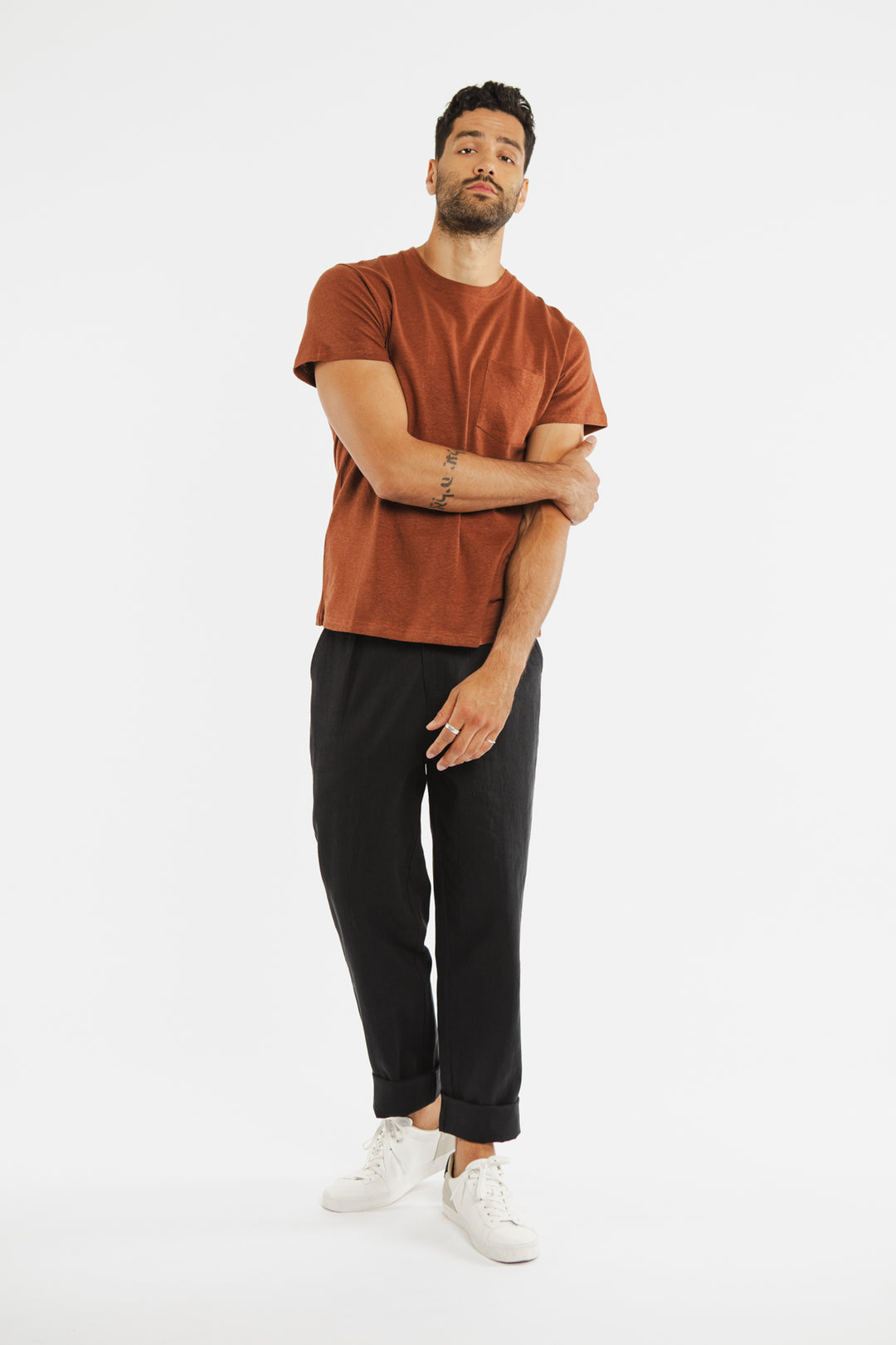A man with short curly hair and a beard stands against a white background wearing the Bridge & Burn Organic Hemp Pocket Tee in Burnt Umber, black pants, and white sneakers. He touches his left arm with his right hand.