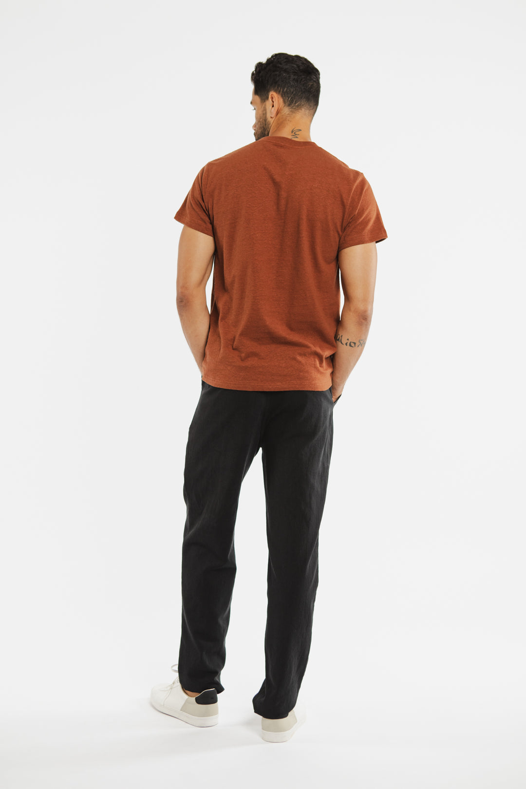 A man faces away from the camera, wearing the Bridge & Burn Organic Hemp Pocket Tee in Burnt Umber, black pants, and white sneakers against a plain white background. His arms are relaxed at his sides.