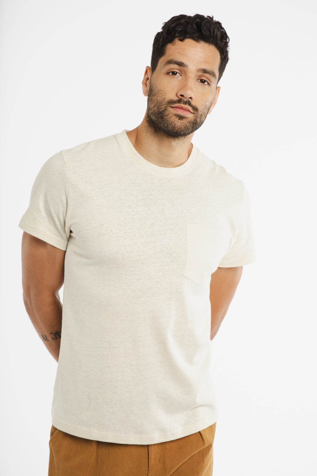 A man with short, dark curly hair and a beard wears the Bridge & Burn Organic Hemp Pocket Tee in ivory and brown pants. He stands against a white background with his hands behind his back, looking slightly to the side.