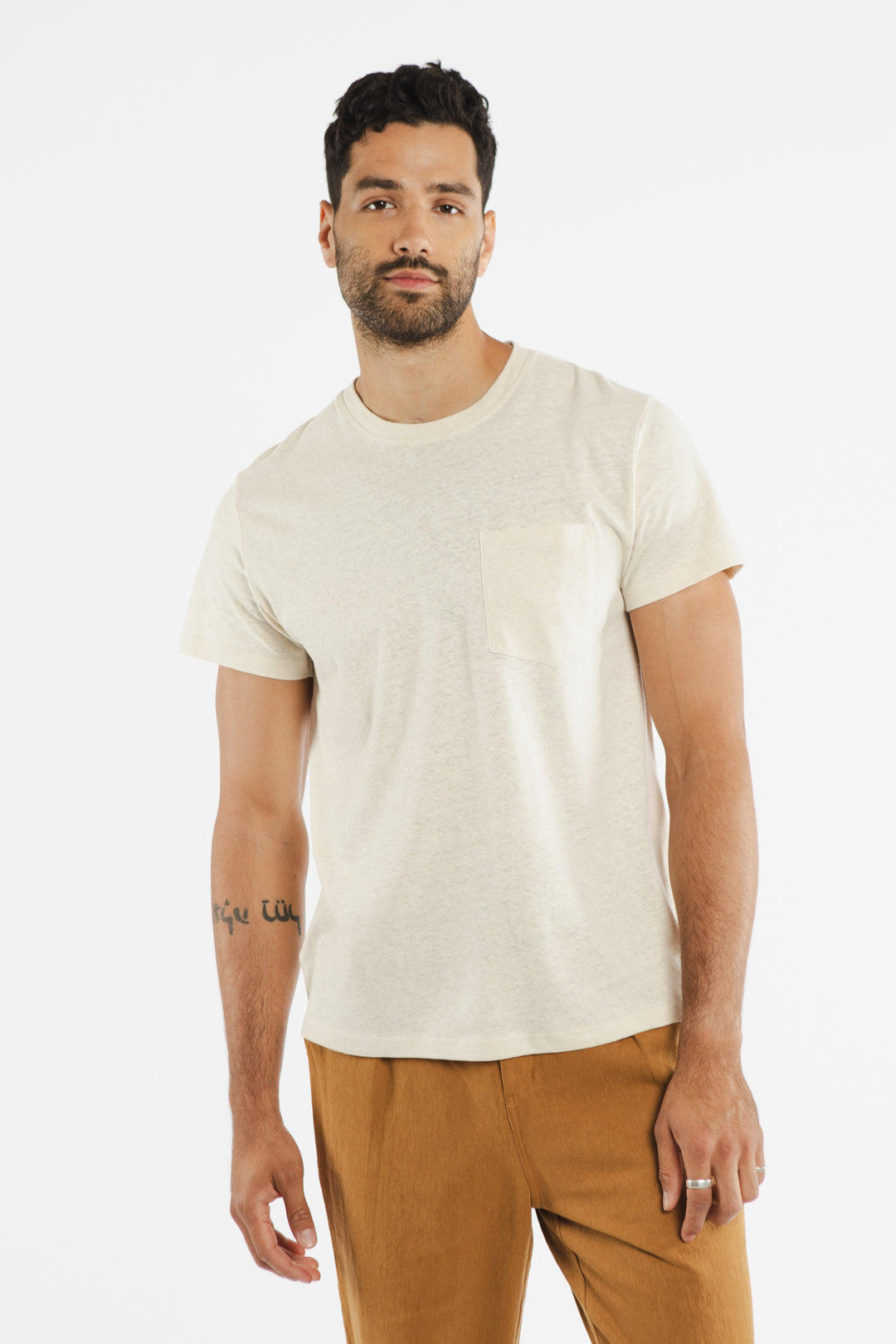 A man with short dark hair, a beard, and a forearm tattoo wears the Bridge & Burn Organic Hemp Pocket Tee in ivory with brown pants, standing against a plain white background.