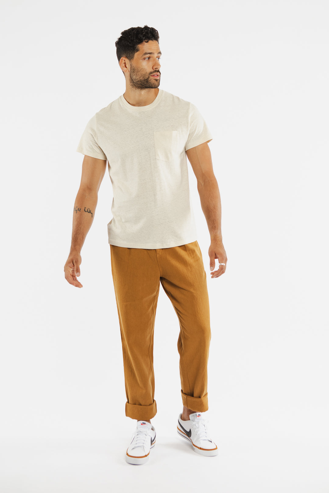 A man with a short beard and curly hair looks to his left, wearing the Bridge & Burn Organic Hemp Pocket Tee in Ivory, mustard pants with rolled cuffs, and white sneakers against a white background.