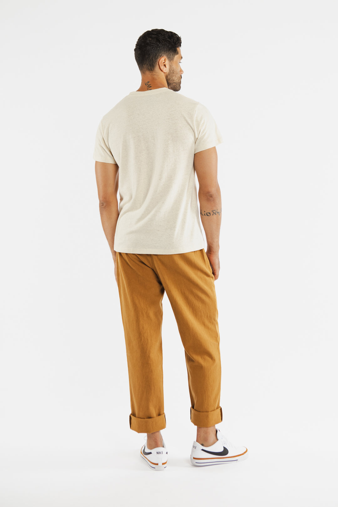 A man faces away from the camera, wearing the Bridge & Burn Organic Hemp Pocket Tee in Ivory. He pairs it with rolled-up brown pants and white Nike sneakers with navy swooshes. The background is plain white.