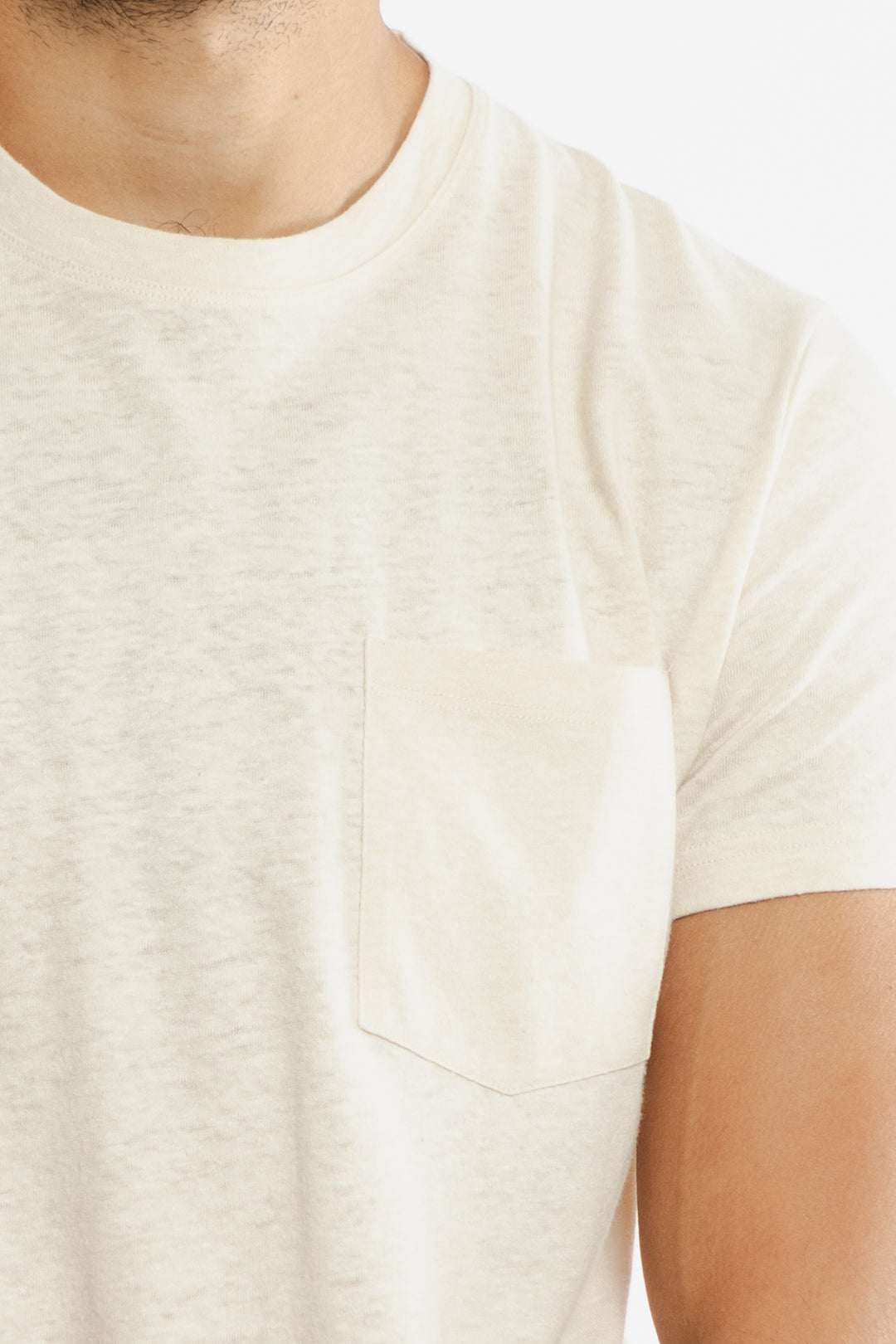 A close-up of a person wearing the Bridge & Burn Organic Hemp Pocket Tee in Ivory, featuring a small chest pocket, with part of their neck, chest, and upper arm visible against a plain background.