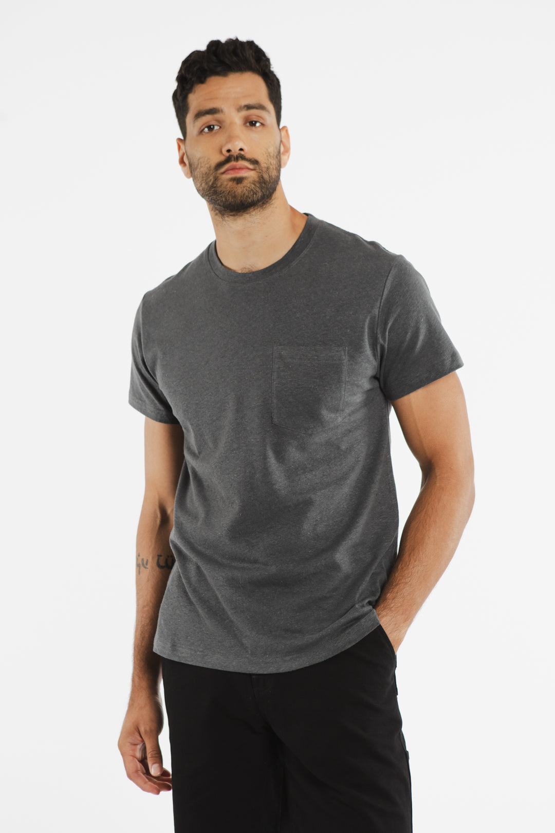 A man with short dark hair and a beard wears the Bridge & Burn Organic Hemp Pocket Tee in Slate Grey with black pants, standing against a plain white background. One hand is in his pocket as he looks calmly at the camera.