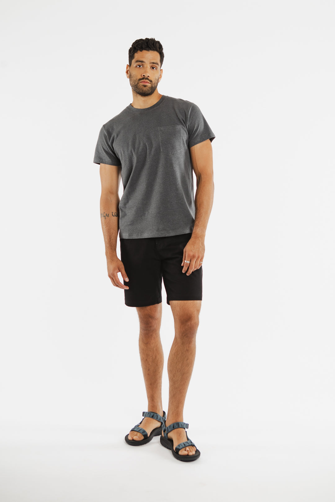 A man with short dark hair and a beard stands against a white background wearing the Bridge & Burn Organic Hemp Pocket Tee in Slate Grey, paired with black shorts and blue sandals. He has a neutral expression, one arm relaxed, the other slightly bent.