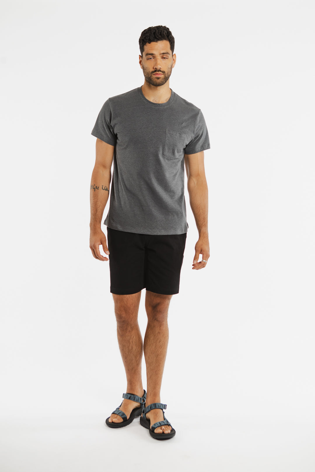 A man with short hair, a beard, and a tattoo on his left forearm stands facing forward against a plain white background, wearing the Bridge & Burn Organic Hemp Pocket Tee in Slate Grey paired with black shorts and blue sandals.