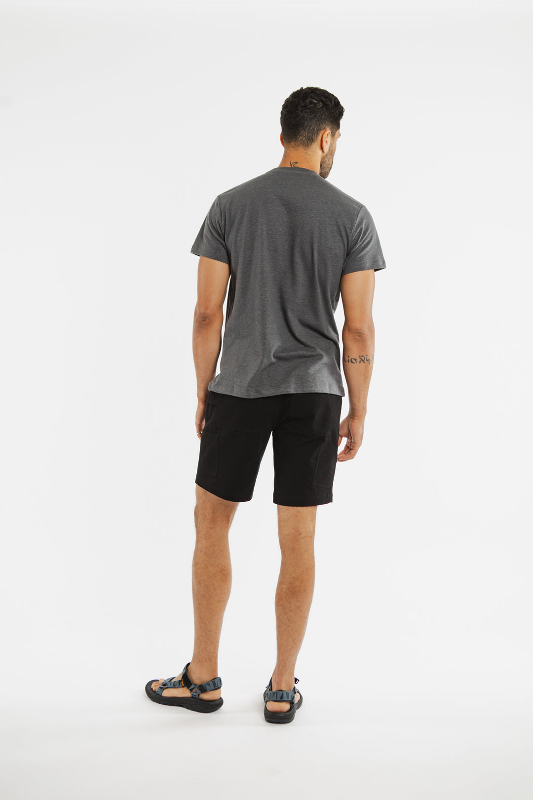 A man stands with his back to the camera, wearing a Bridge & Burn Organic Hemp Pocket Tee in Slate Grey, paired with black shorts and sandals against a plain white background. His arms hang relaxed at his sides.