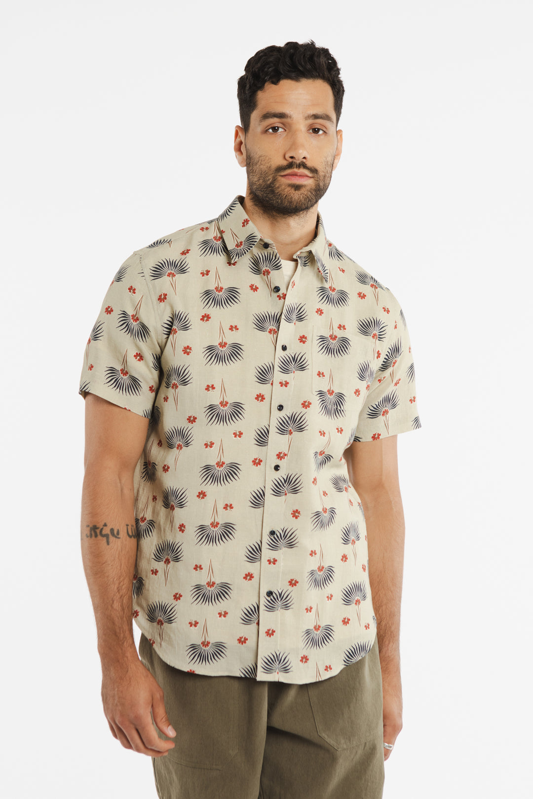 A man with short dark hair and a beard is wearing the Bridge & Burn Harbor Slim Shirt in Aloha Floral—a beige, short-sleeve slim fit shirt featuring a red and black pattern, crafted from a comfortable linen-cotton blend—paired with olive green pants, standing against a plain white background.